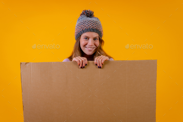 Smiling woman in wool cap pointing at cardboard sign on yellow